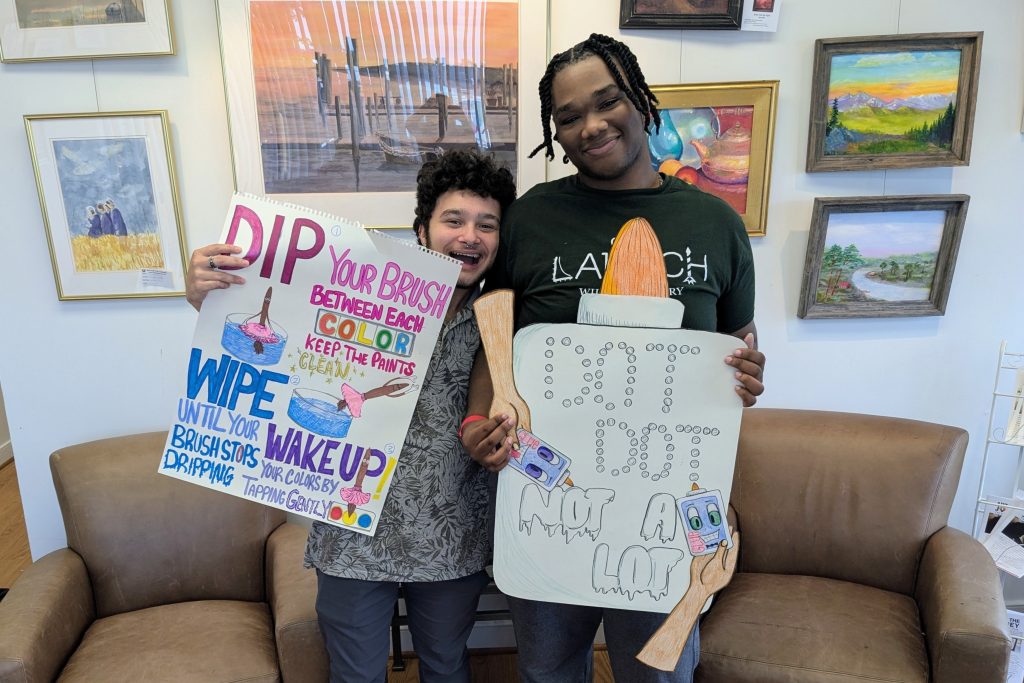 Two young men pose in front of a gallery wall at the AFJ studio holding posters they made for the art classroom